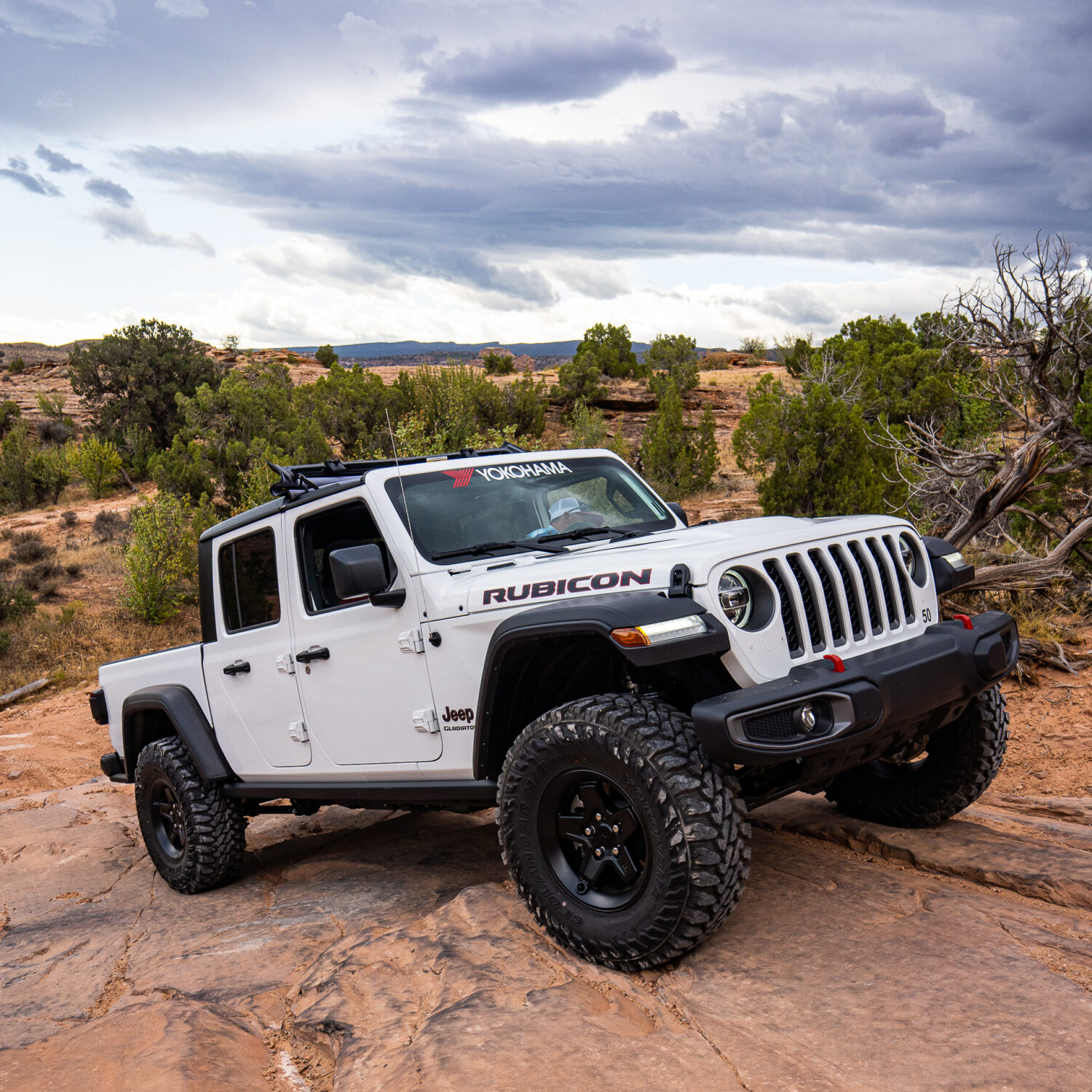 White Jeep Rubicon with Yokohama branding on rocky terrain under a cloudy sky.