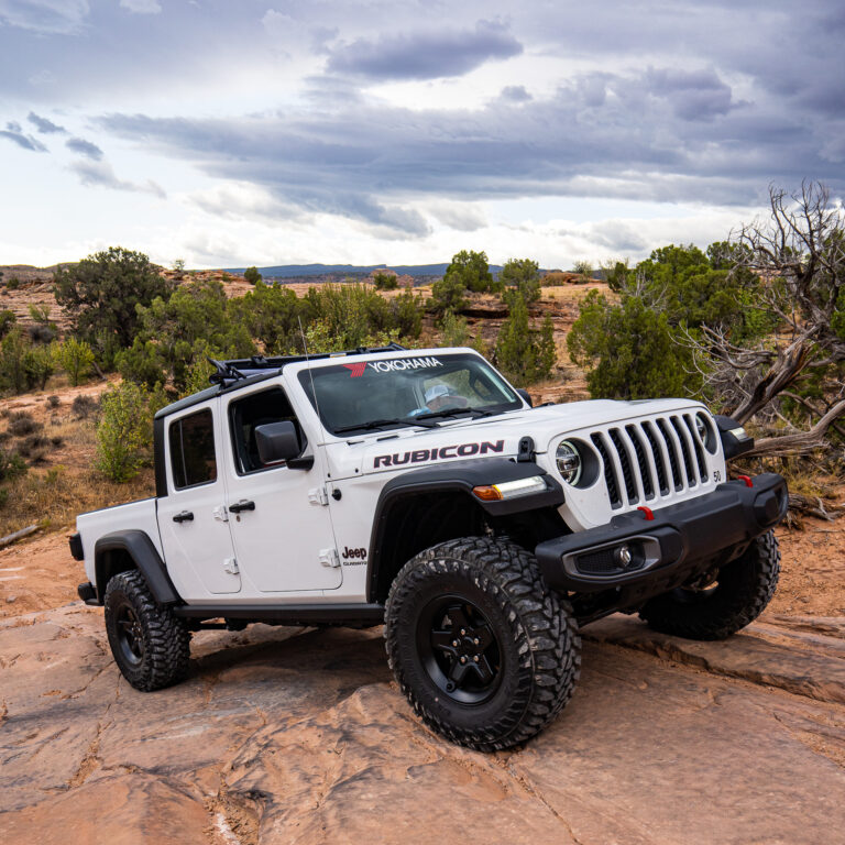 White Jeep Rubicon with Yokohama branding on rocky terrain under a cloudy sky.
